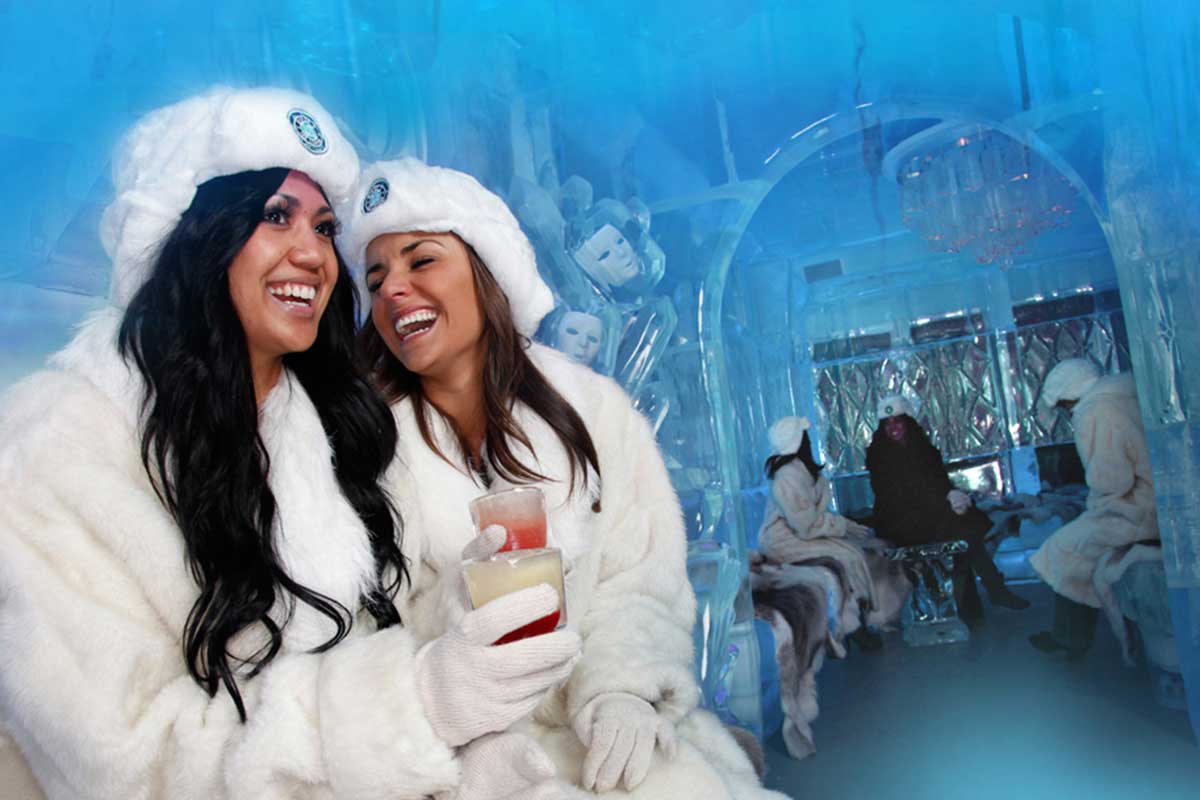 Two women laughing, dressed in white fur coats and hats, holding drinks in an ice bar with more people in the background.