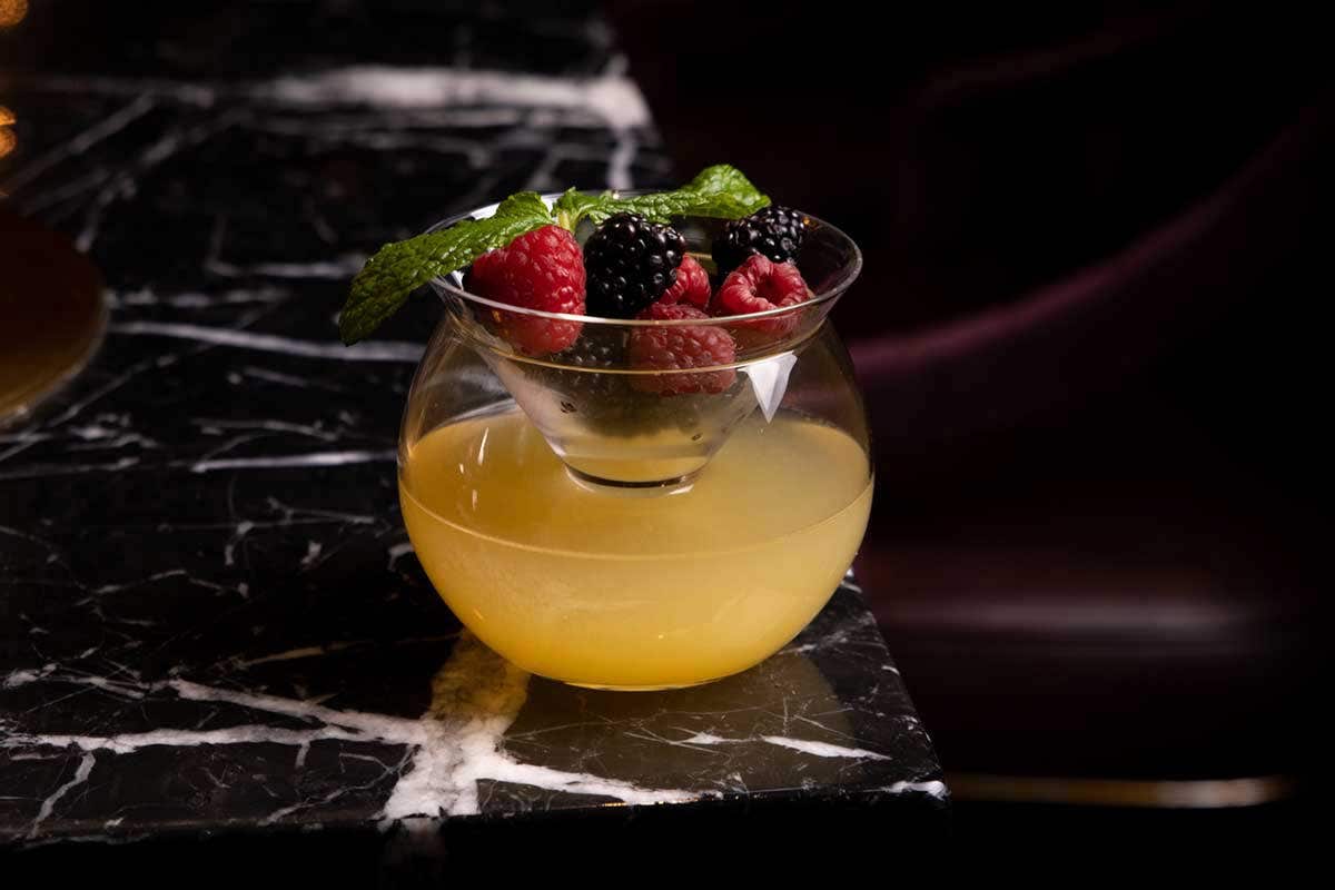 A cocktail in a round glass with berries and mint on top, placed on a black marble surface.