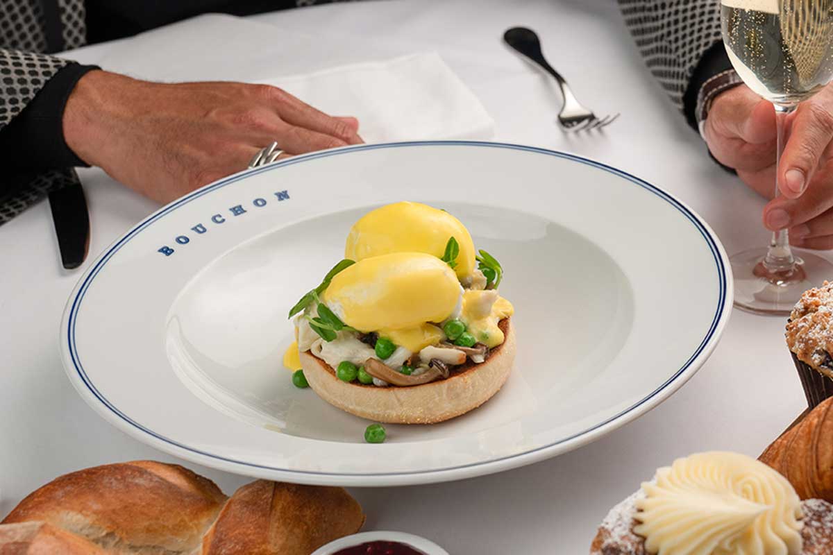 Two poached eggs with hollandaise sauce on a muffin, illustrated in a Bouchon plate, with bread and a glass of champagne.