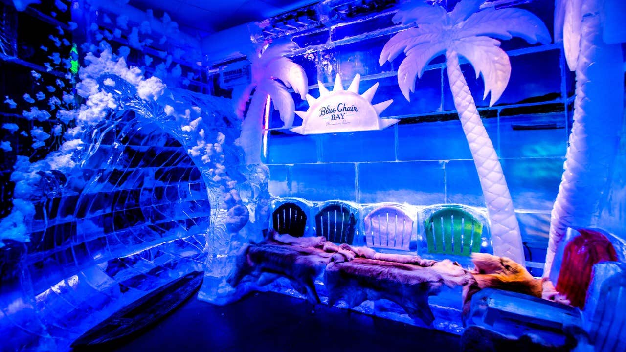 Colorful chairs, palm trees, and ice sculptures in a blue-lit ice bar with a "Blue Chair Bay" sign above the seating area.