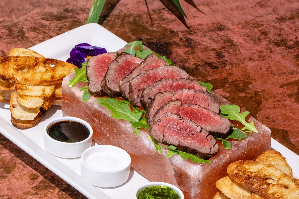 Slices of rare steak served on a Himalayan salt block with toasted bread and dipping sauces on a white rectangular plate.