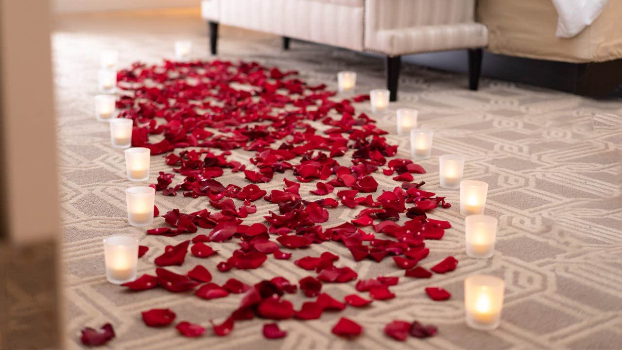 A bed with a pathway of scattered red rose petals and lit candles leading towards it on a patterned carpeted floor.