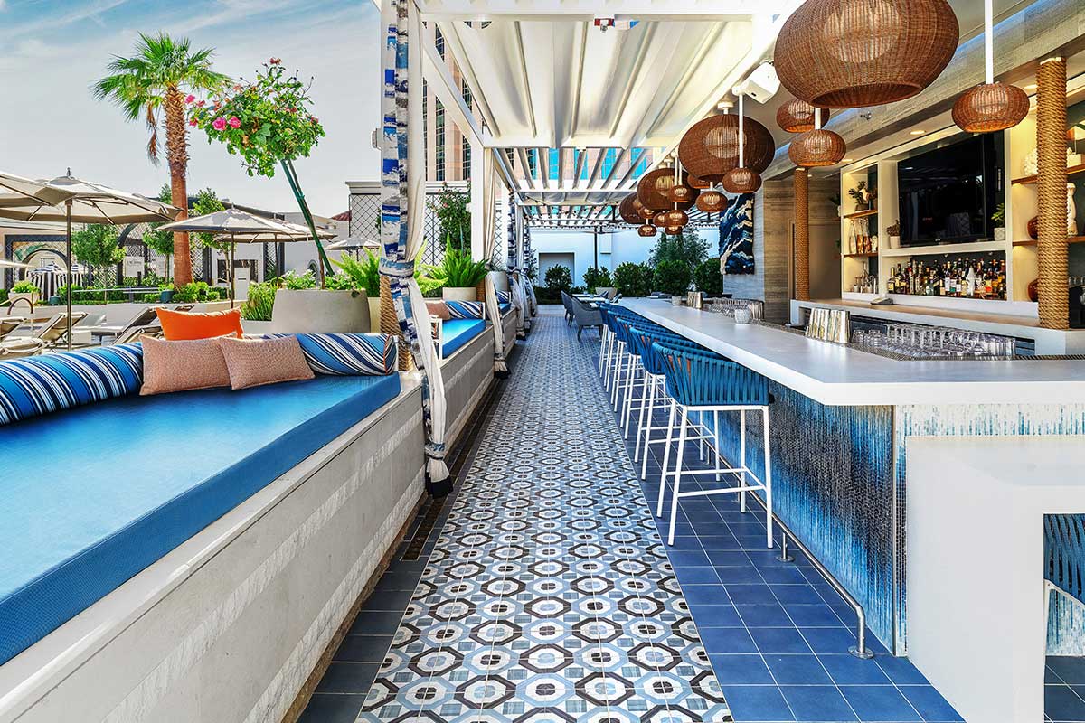 A stylish outdoor bar with modern seating, decorative floor tiles, hanging wicker lights, and a distant palm tree.