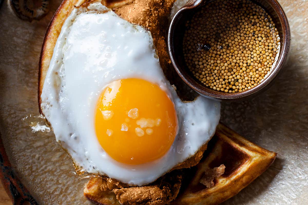 Sunny-side up egg on a waffle with a small bowl of mustard seeds on the side.