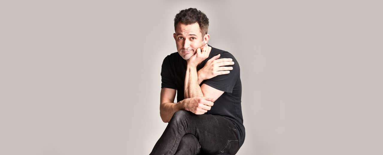 A man in a black shirt and jeans sits on a stool, looking at the camera with his arms resting on his knees.