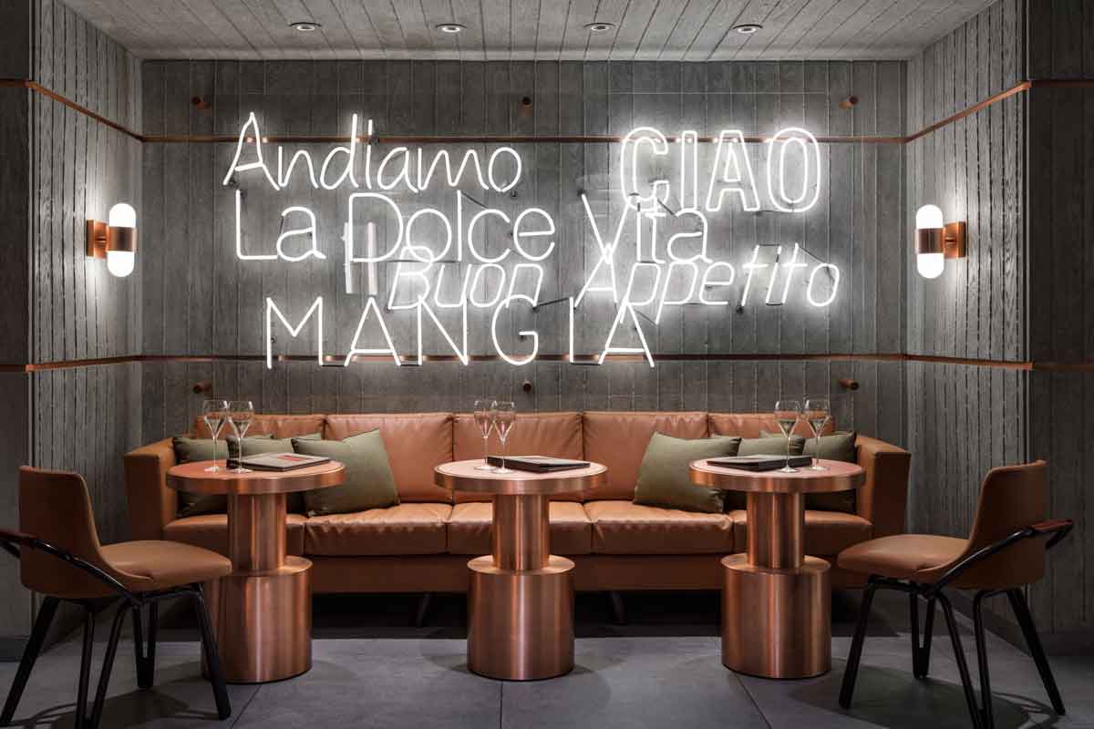 Trendy cafe with modern decor, neon signs reading Italian phrases, round tables, and a brown sofa.