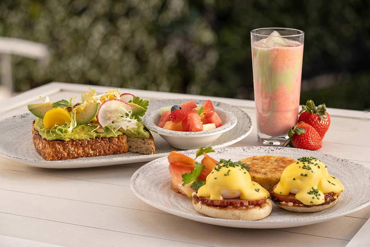 A breakfast spread including avocado toast, eggs Benedict, fruit salad, a smoothie, and fresh strawberries.