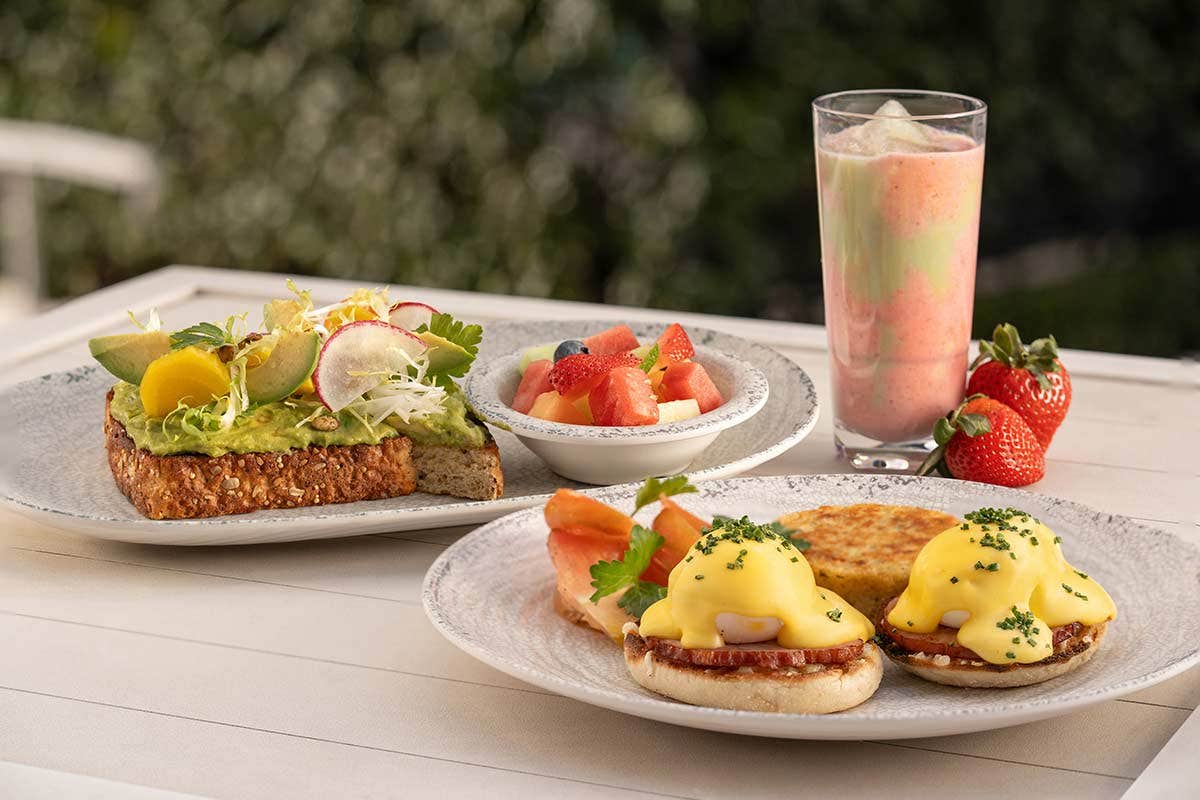 A breakfast spread including avocado toast, eggs Benedict, fruit salad, a smoothie, and fresh strawberries.