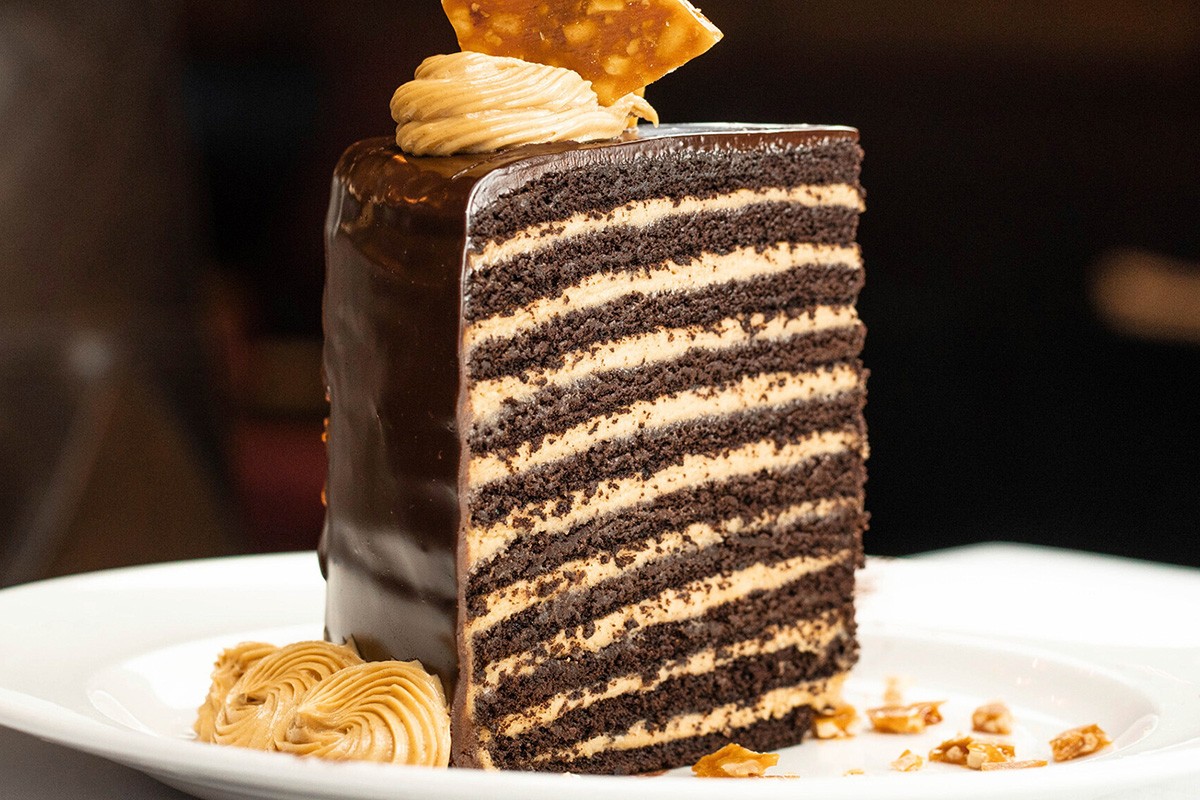 A piece of layered chocolate and peanut butter cake with a frosting swirl and toffee garnish on a white plate.