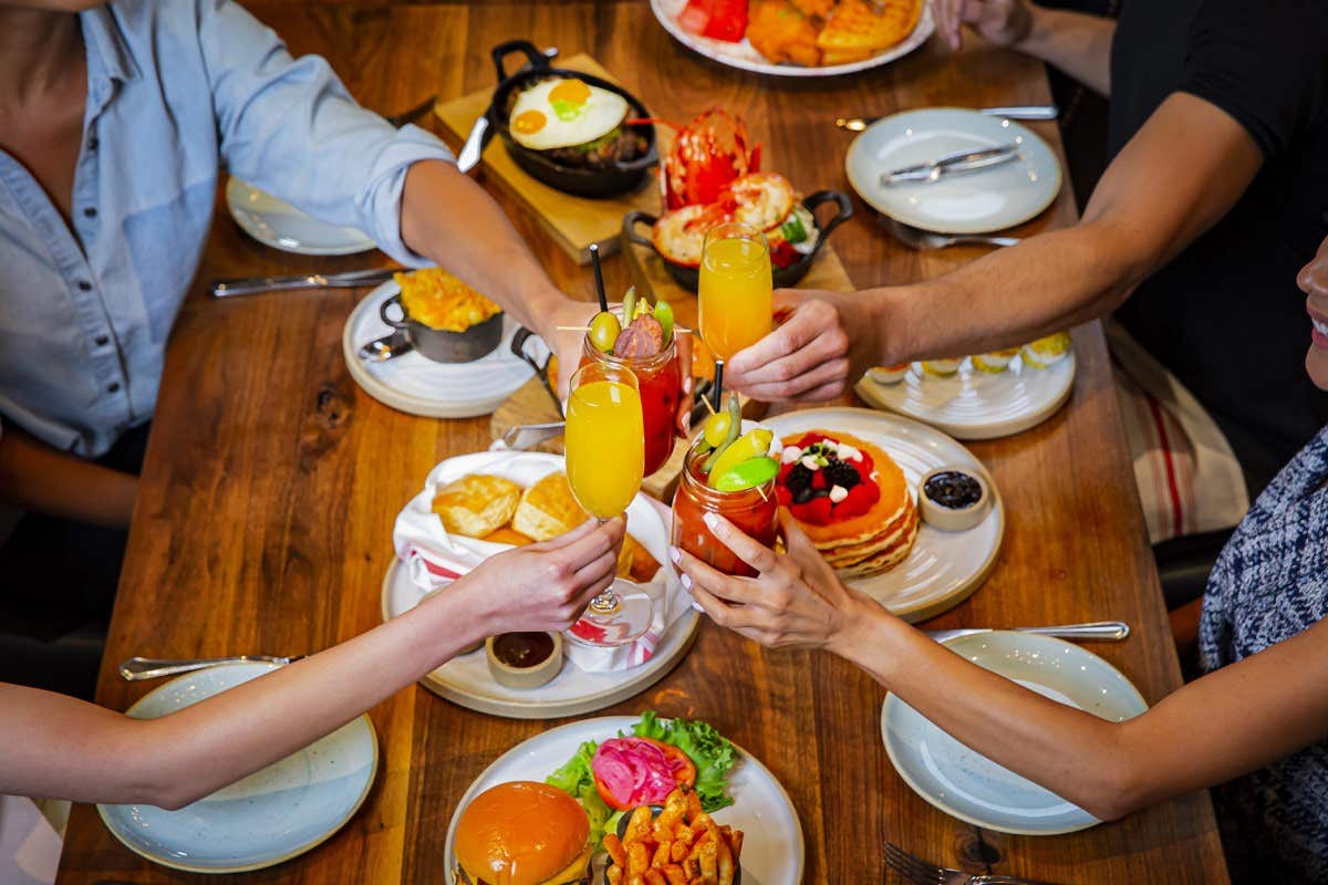 Four people clinking glasses over a table filled with breakfast foods, including pancakes and eggs.