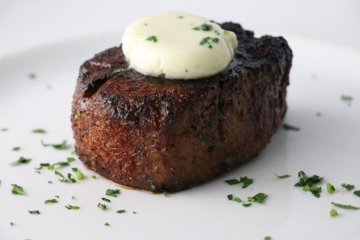 A juicy, grilled steak topped with a pat of melting butter and garnished with chopped herbs on a white plate.