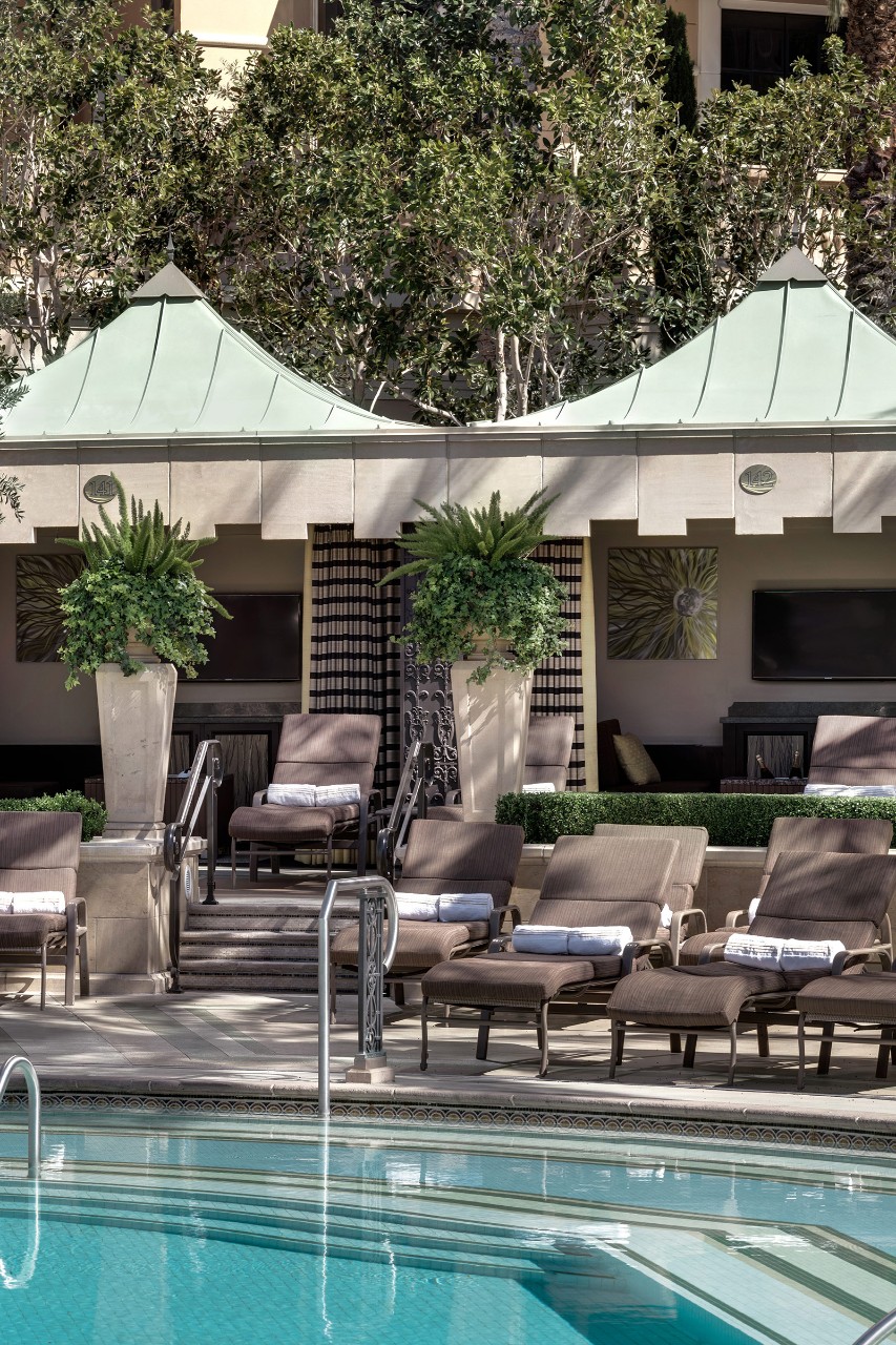 Poolside cabanas with lounge chairs and potted plants beside a clear blue swimming pool.