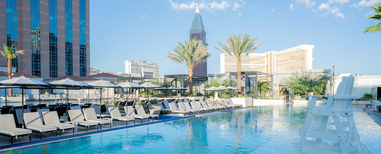 Luxurious hotel pool area with lounge chairs, umbrellas, and a backdrop of tall buildings under a clear blue sky.