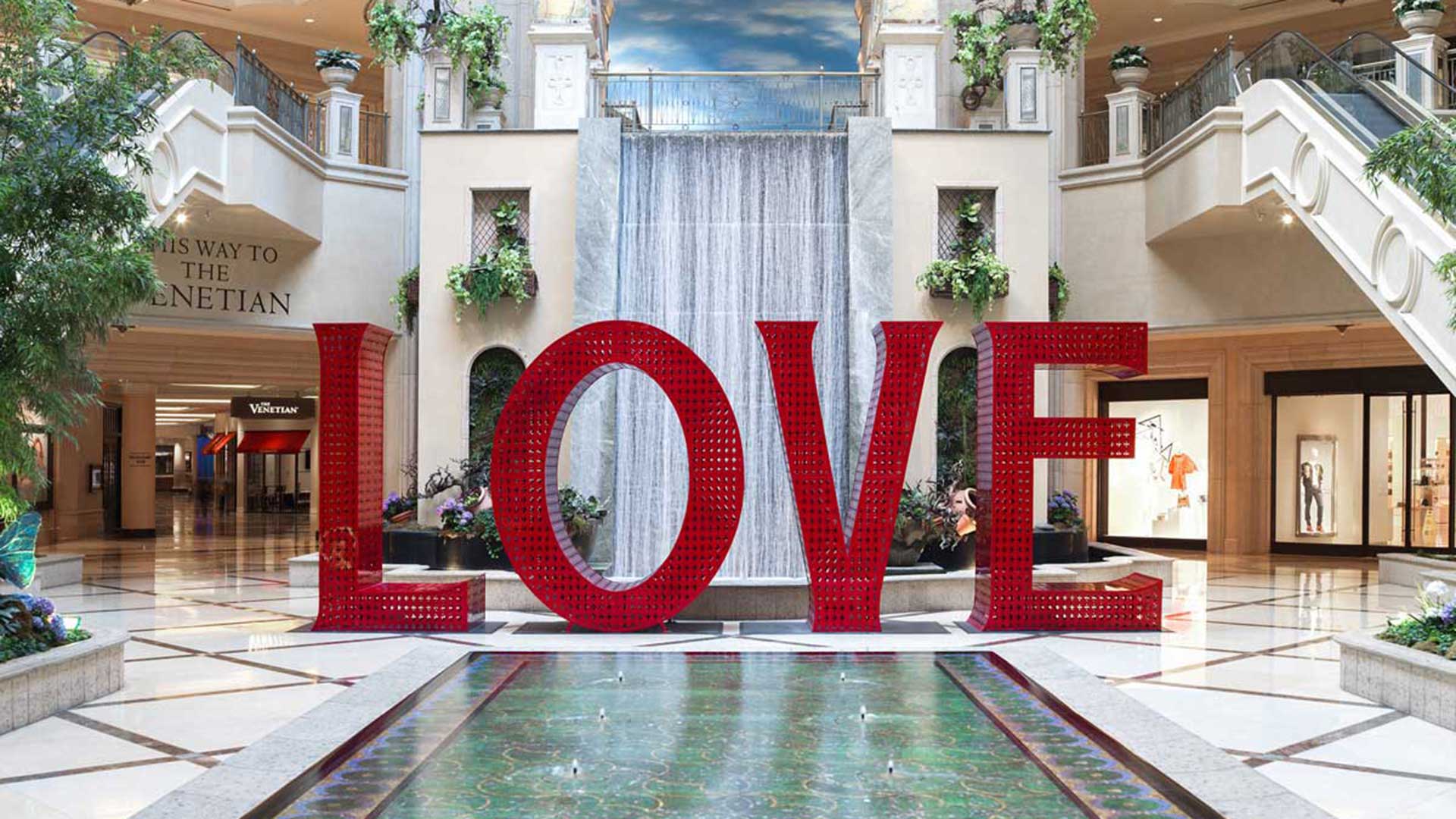 Large red "LOVE" sculpture with a waterfall backdrop and lush greenery in a spacious, elegant indoor setting.