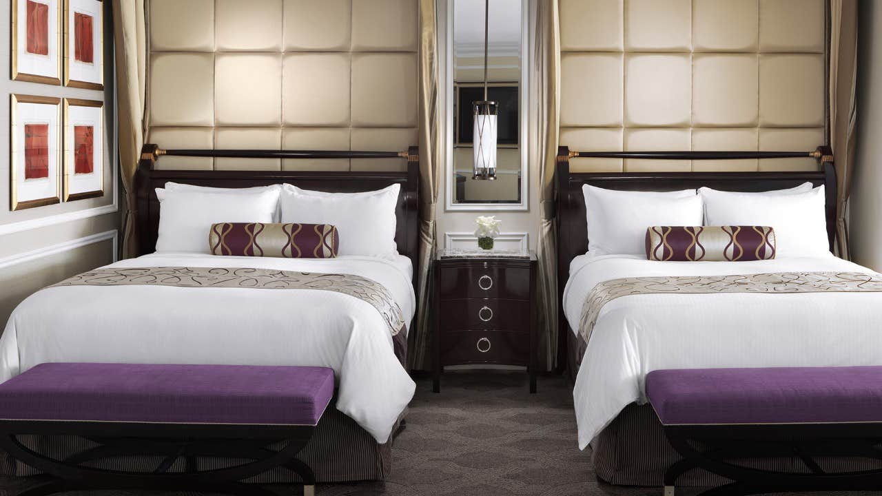 A hotel room with two double beds, white bedding, purple benches, and a nightstand with a flower vase in between.