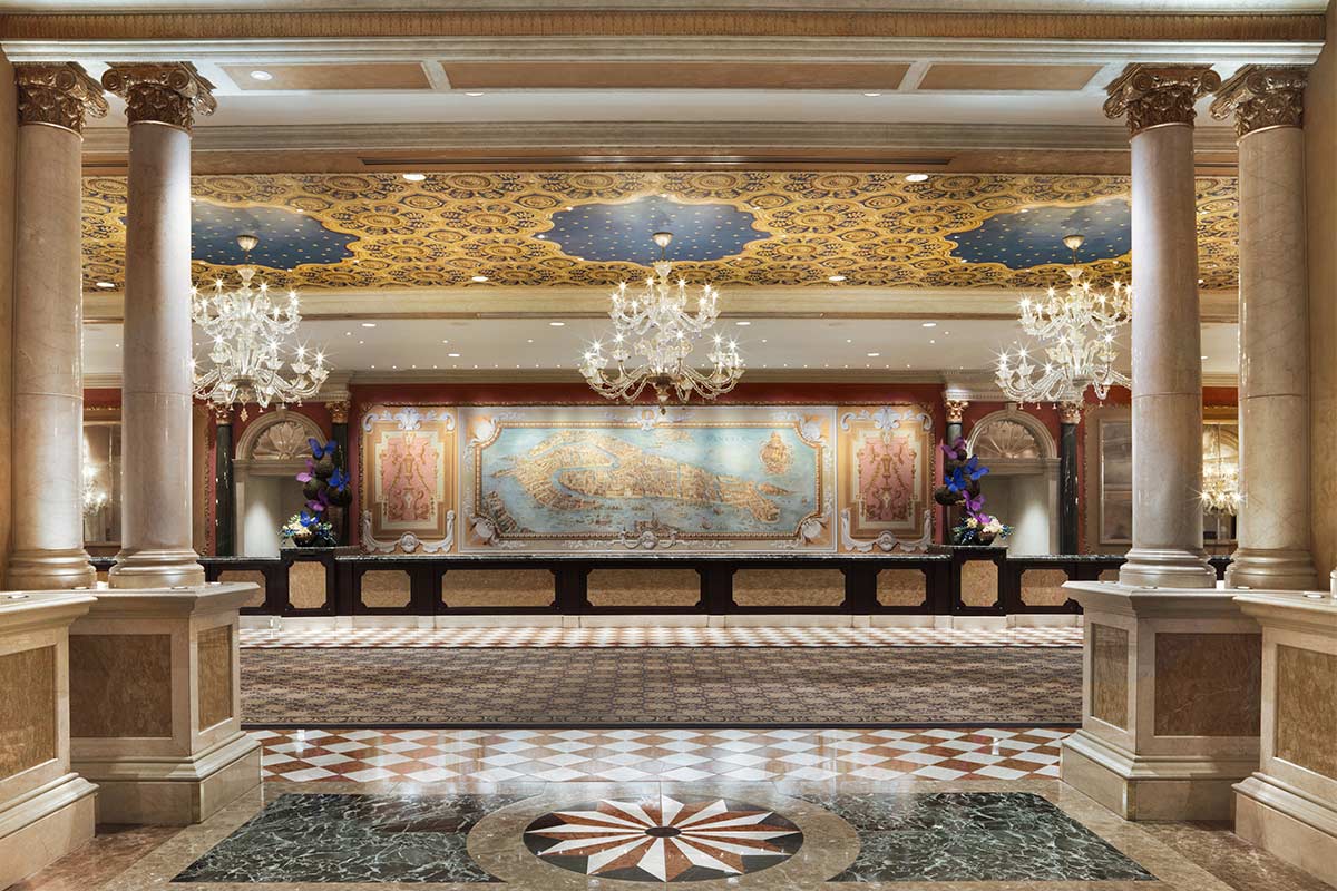 Elegant hotel lobby with ornate ceilings, chandeliers, marble columns, and a large mosaic artwork behind the reception desk.