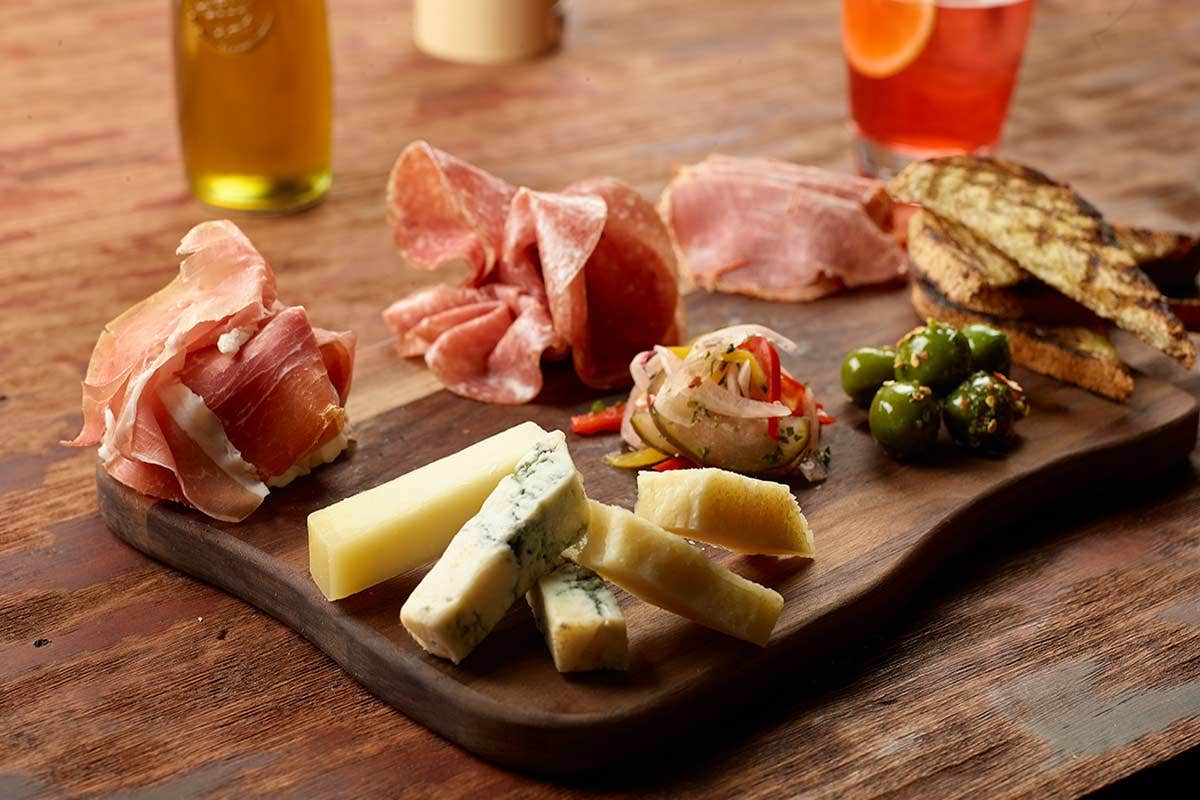 A charcuterie board with cured meats, cheeses, olives, pickled vegetables, and toasted bread on a wooden table with drinks.