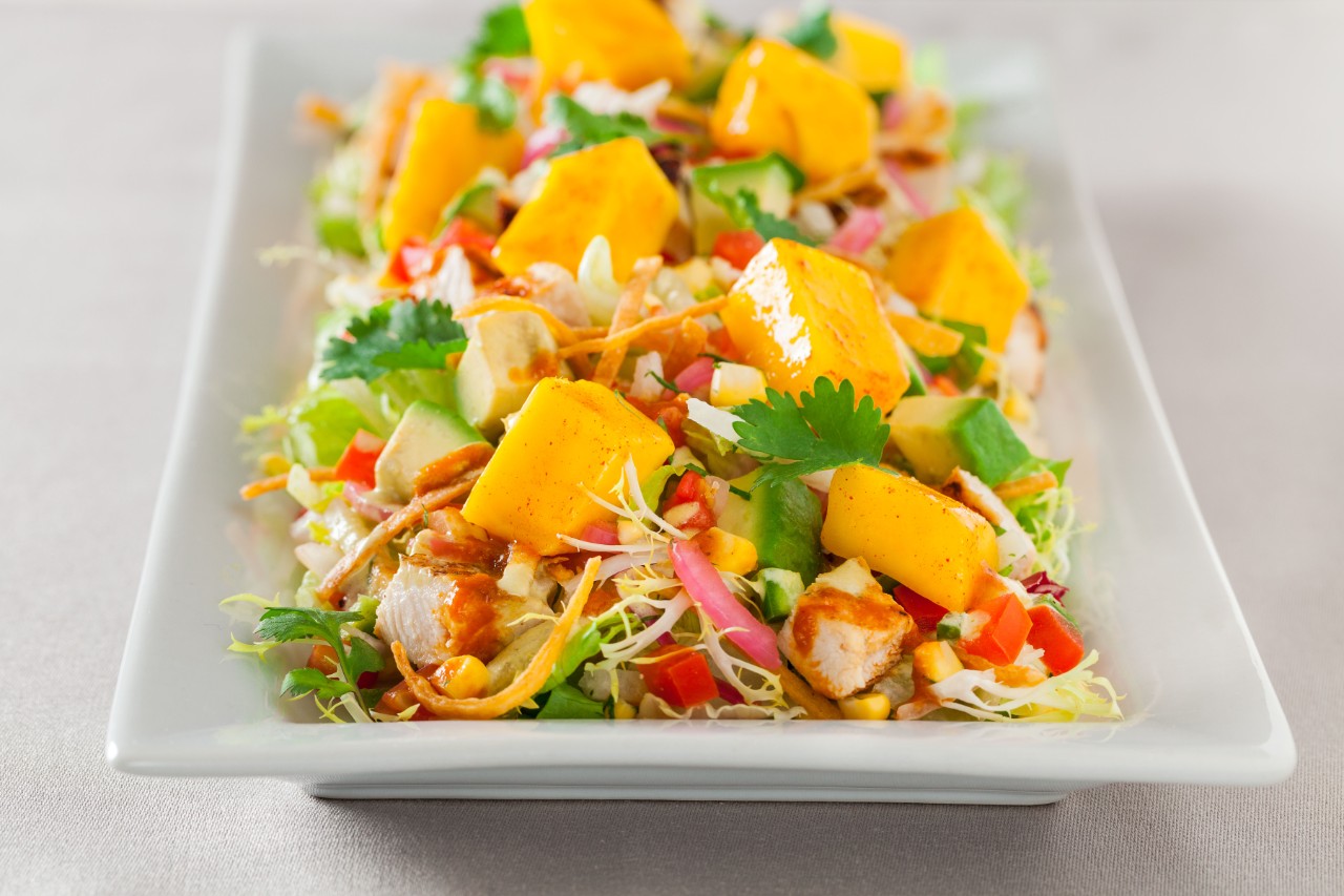 A vibrant mango chicken salad with cilantro, mixed greens, and vegetables on a white rectangular plate.