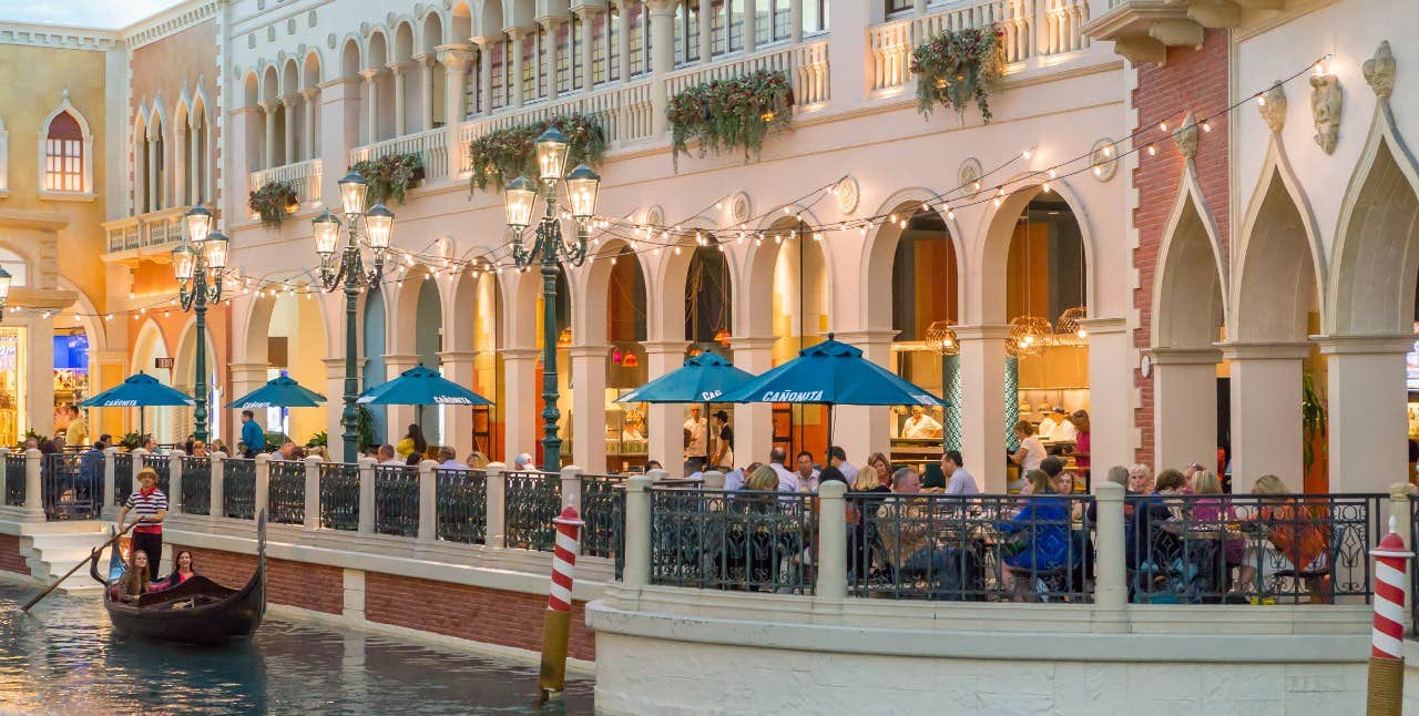 People enjoy outdoor dining next to a canal with a gondola in a plaza designed to resemble Venice.