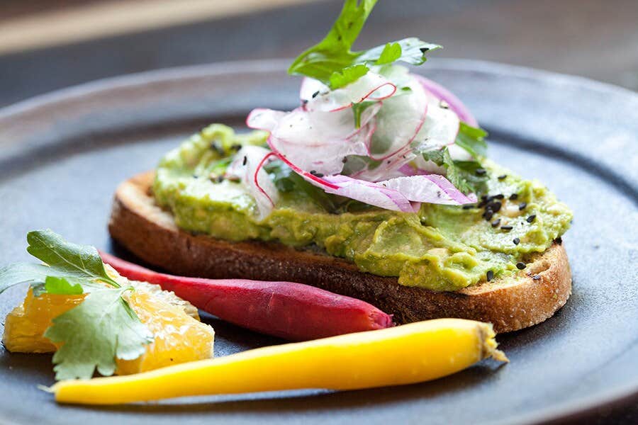 Avocado toast topped with sliced radishes and greens, served with colorful carrots and an orange wedge on a dark plate.