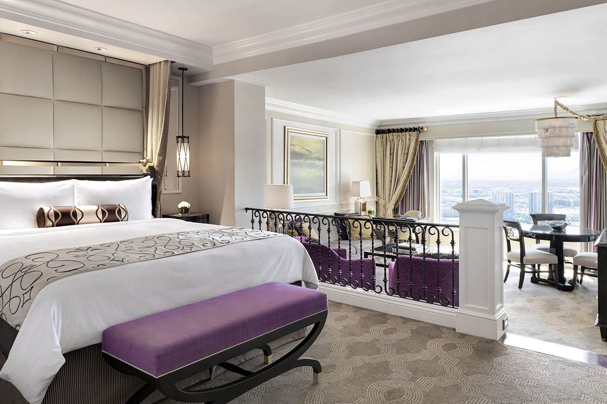 Elegant hotel room with a king-sized bed, seating area, purple accents, and a scenic city view through large windows.