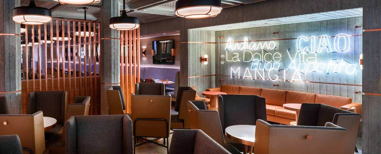 Modern restaurant interior with neon signs and cozy seating, including armchairs and round tables. Wooden partitions divide spaces.