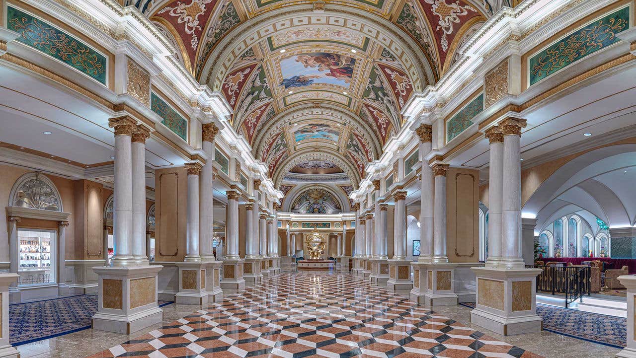 A grand, ornate hallway with tall marble columns, detailed ceiling murals, and a central gold statue at the end.