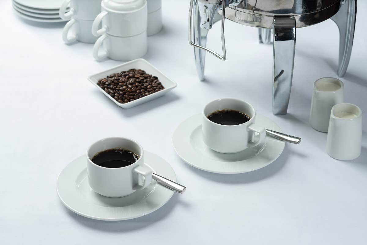 Two white coffee cups on saucers, a plate of coffee beans, a coffee urn, and milk pitchers on a white tablecloth.