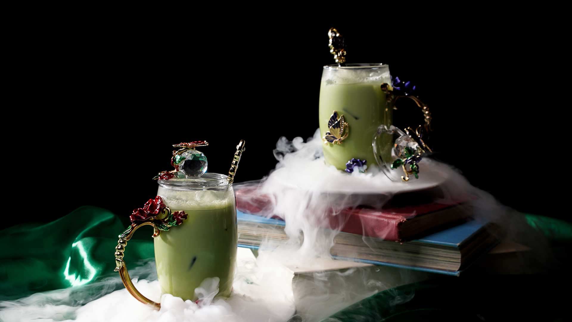 Two ornate glasses of green beverage with mist around them, placed on books against a dark background.