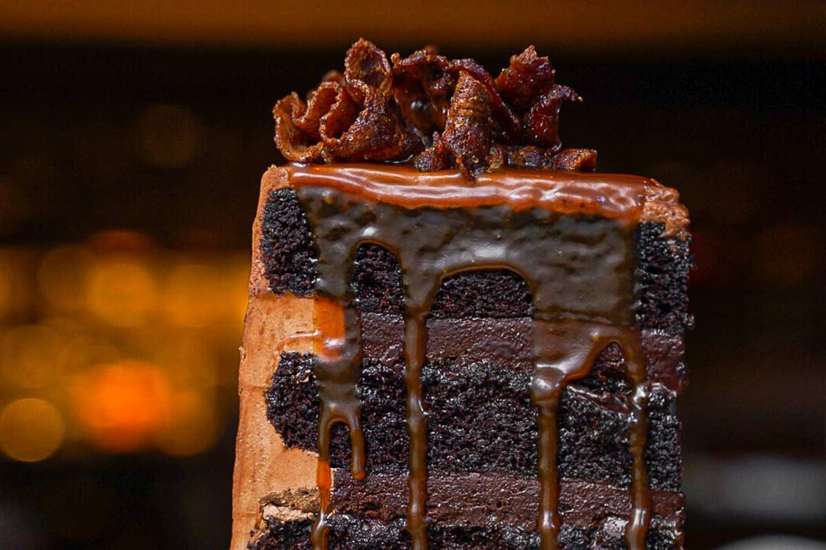 A close-up of a rich chocolate cake with caramel drizzle and bacon strips on top.