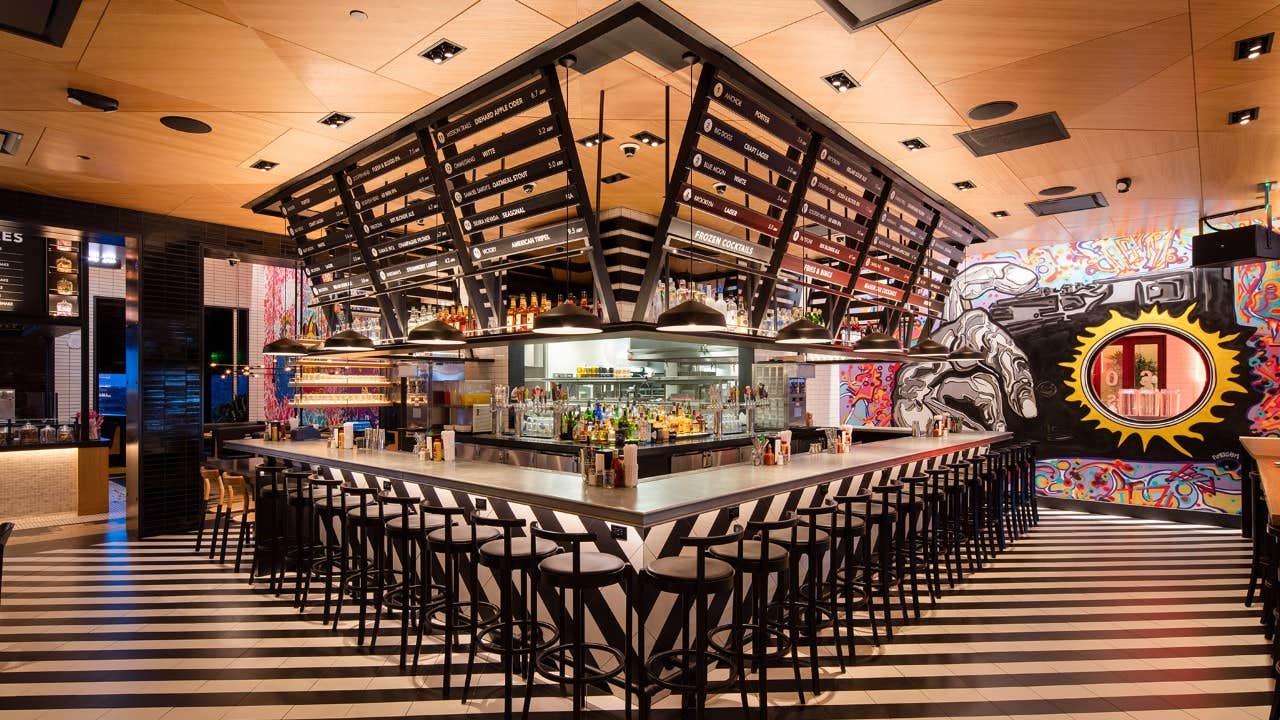 A modern bar with black stools, overhead shelves, colorful murals, and checkered flooring inside a vibrant venue.