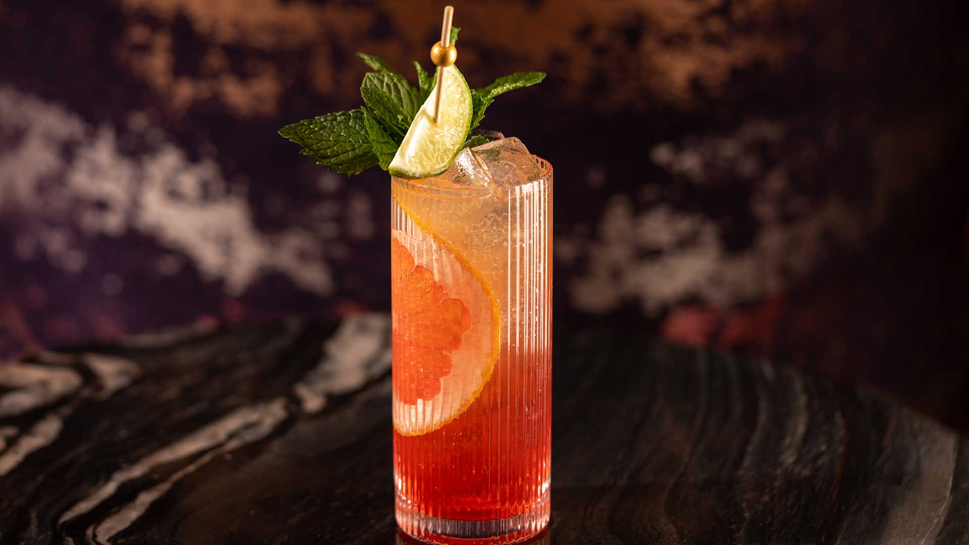 A tall glass with a red-orange cocktail garnished with lime, mint, and a slice of grapefruit on a dark marble table.