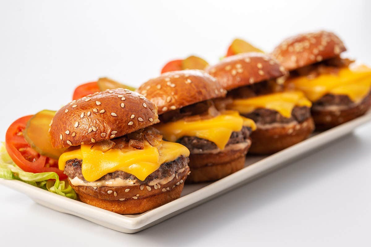 A plate of four cheeseburger sliders with sesame seed buns, topped with cheese, pickles, tomatoes, and lettuce.