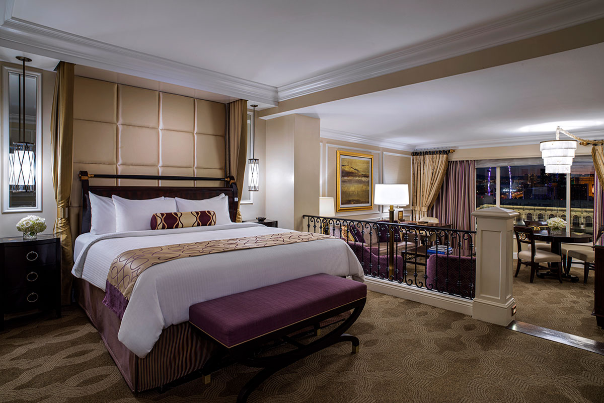 Luxurious hotel suite with a king-sized bed, a sitting area, elegant decor, a large window, and ambient lighting.