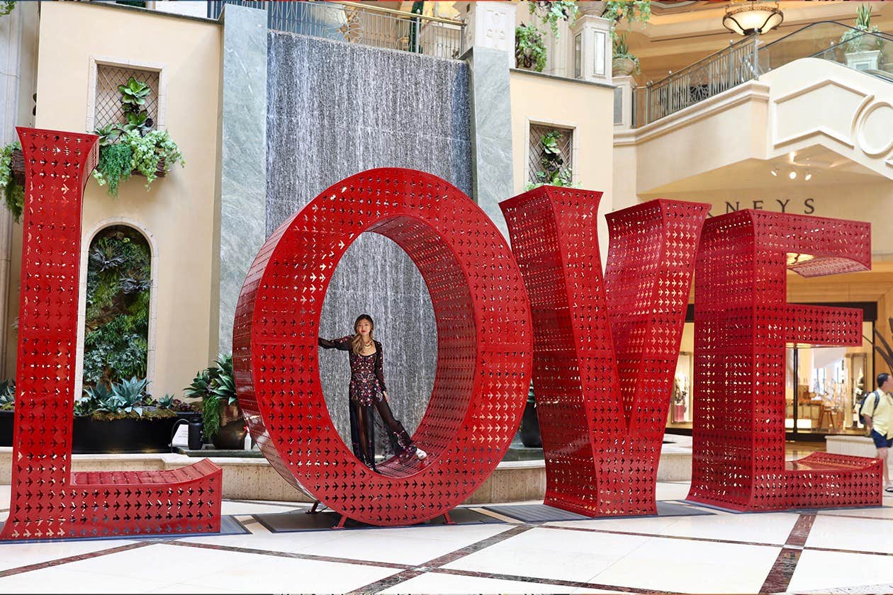 A person stands inside the letter "O" in a large, red "LOVE" sculpture in an indoor setting with greenery and a waterfall.