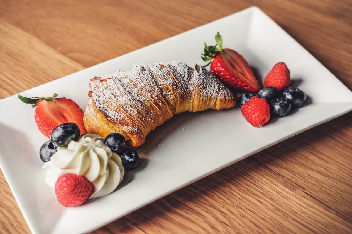 A croissant topped with powdered sugar, served with strawberries, blueberries, raspberries, and a dollop of whipped cream.