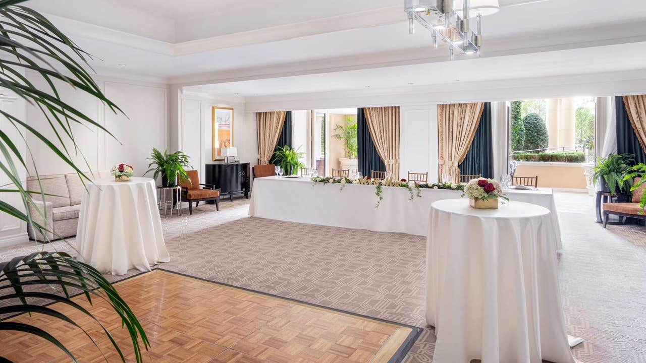 Bright and elegant reception room with round tables, floral arrangements, chairs, large windows, and a chandelier.