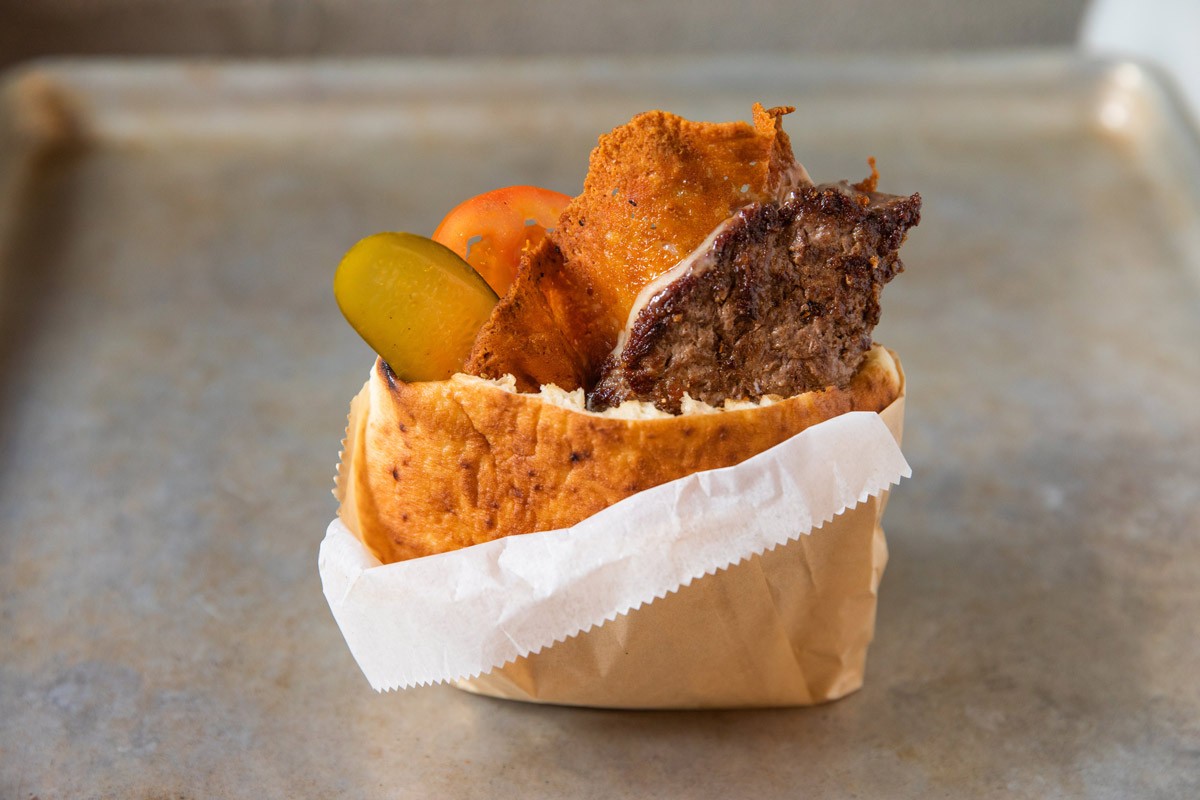 A sandwich with roasted meat, pickles, and tomato is wrapped in paper on a metal tray.