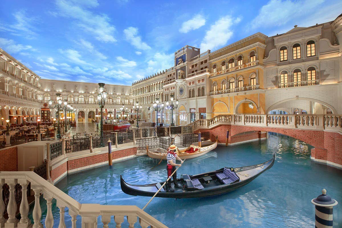 Indoor canal scene with gondola and gondolier, surrounded by Venetian-style buildings under a painted blue sky.