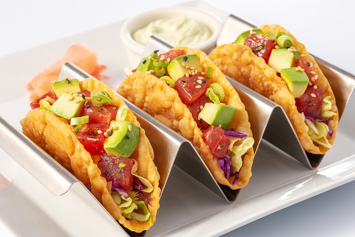 Three crispy tacos filled with diced avocado, tuna, and vegetables, served on a metal stand with a side of dipping sauce.