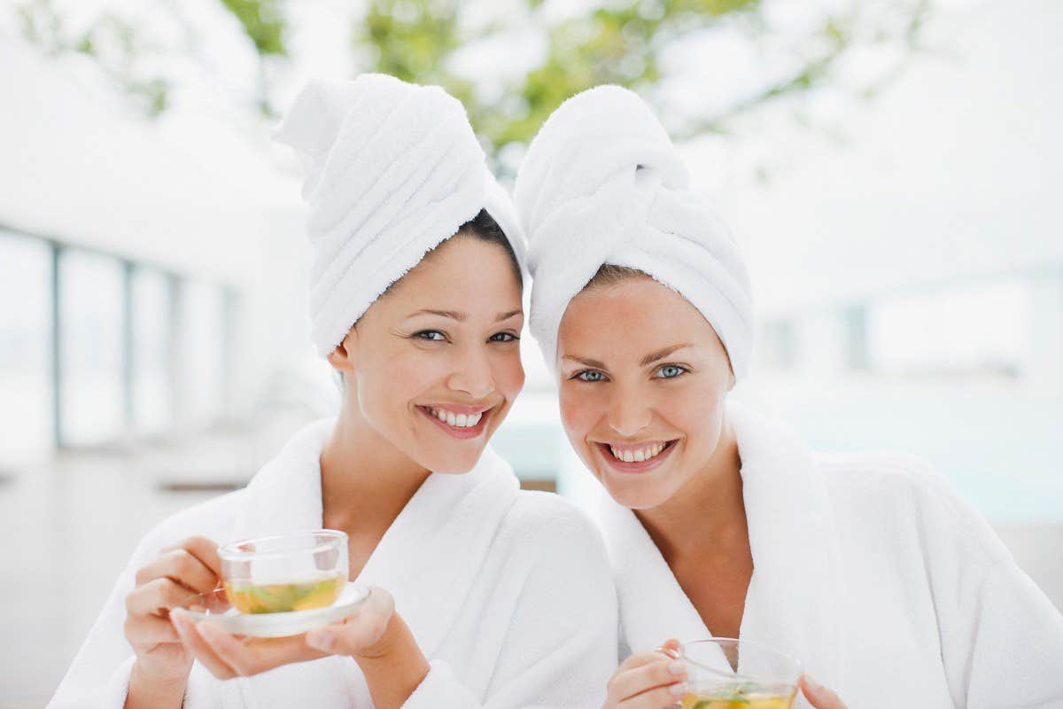 Two smiling women in white robes and towels on their heads, holding cups of tea at a spa.