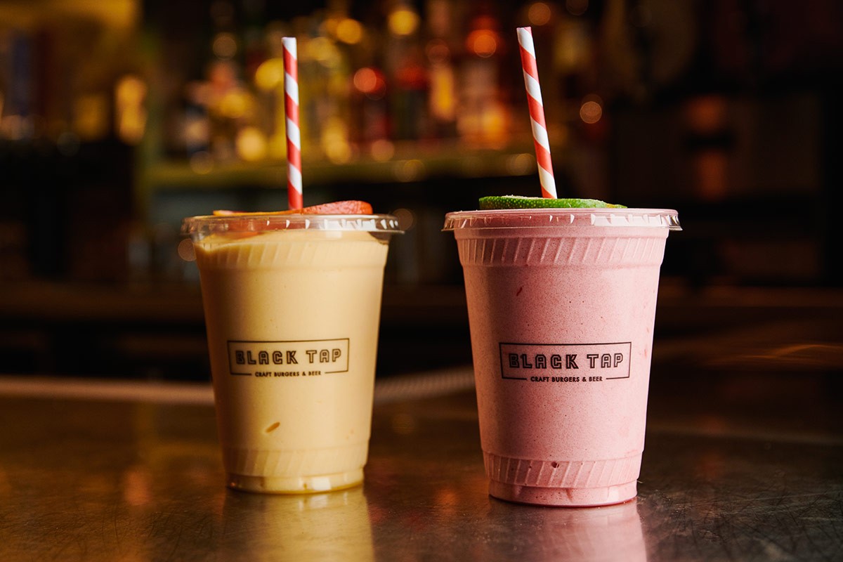 Two milkshakes in plastic cups with red and white striped straws on a counter. One is yellow, and the other is pink.