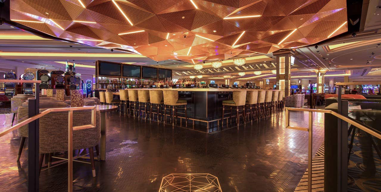 Modern casino bar with geometric ceiling lights, surrounded by bar stools and gaming machines in the background.