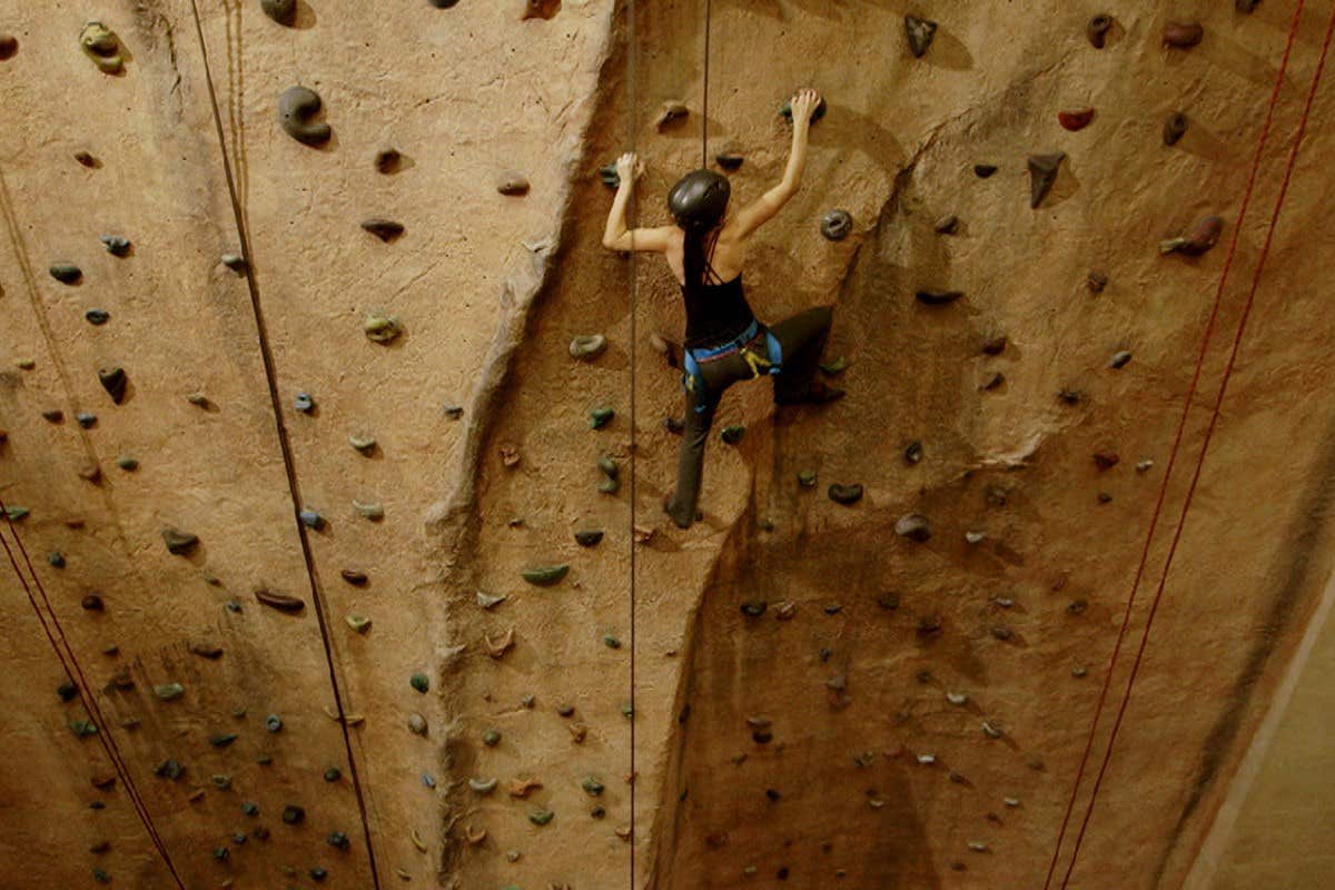 Person rock climbing indoors with a safety harness, helmet, and climbing shoes, scaling a textured wall with holds.