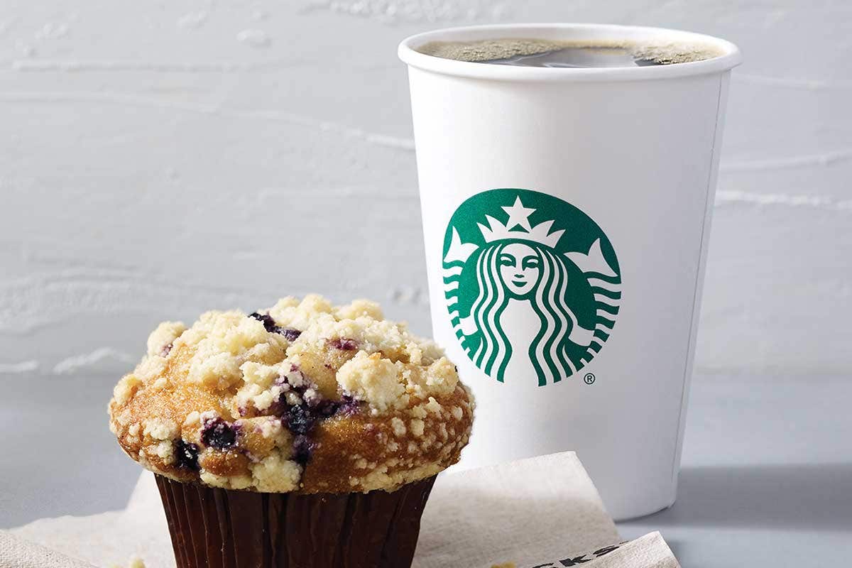 A Starbucks cup of coffee sits next to a blueberry muffin topped with crumbly streusel on a napkin.