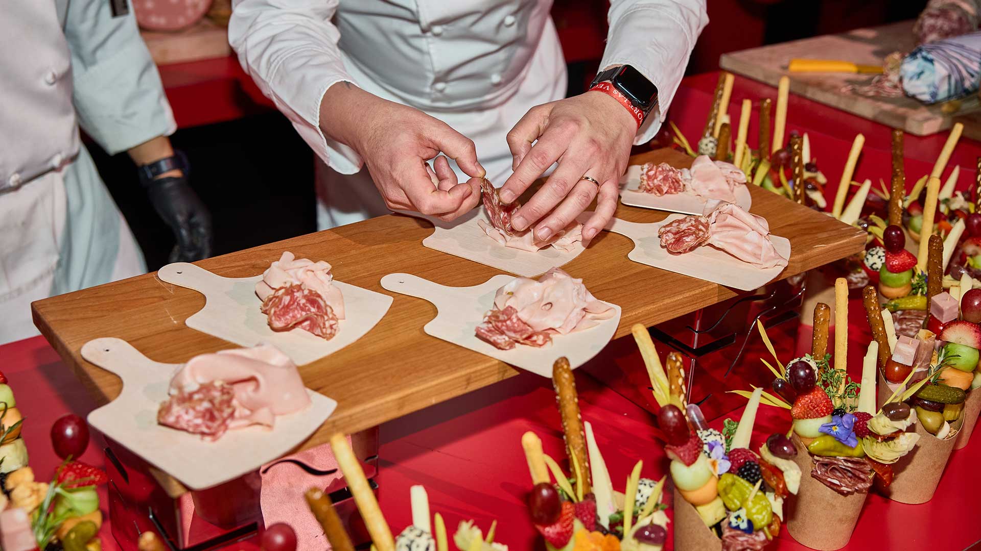 A chef assembles charcuterie boards, arranging cured meats, among an array of assorted snacks and fruit skewers.