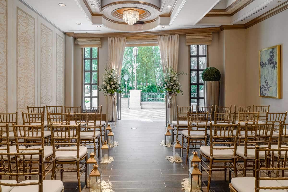 Elegant indoor wedding venue with rows of gold chairs facing a decorated archway, framed by large windows and floral arrangements.