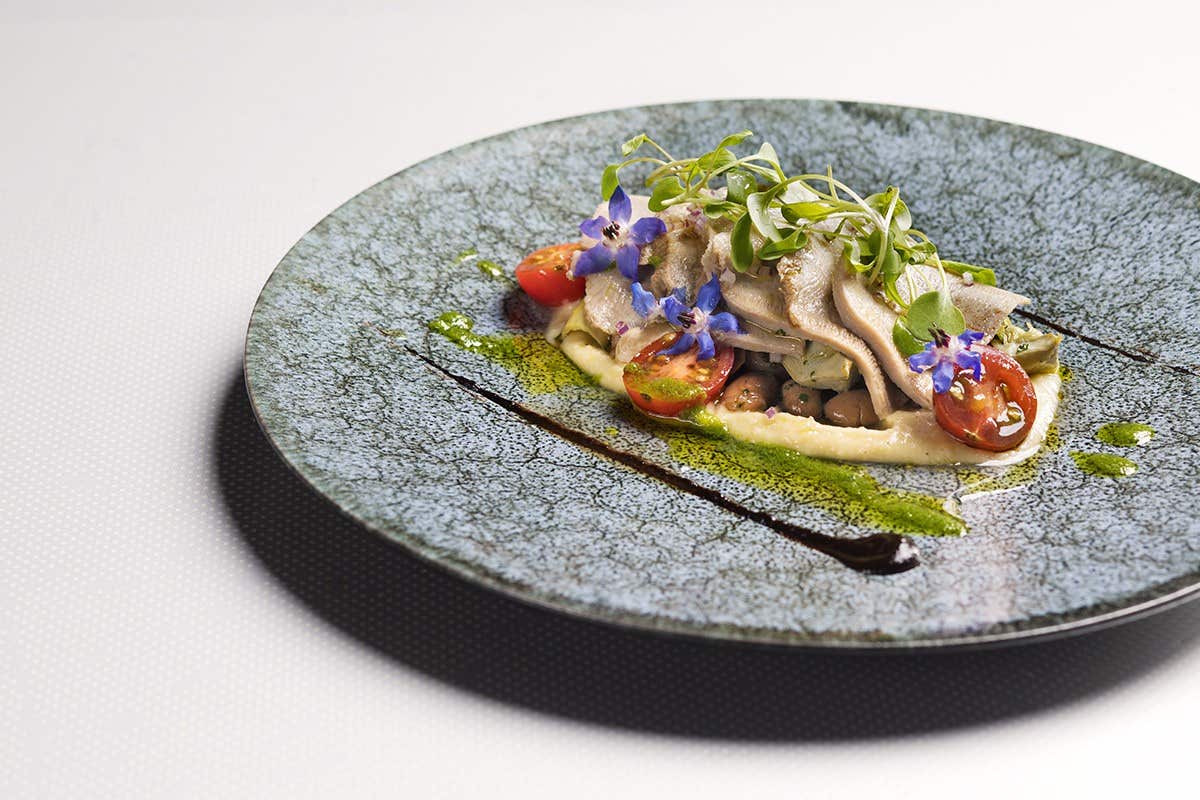 A gourmet dish arranged on a speckled blue plate, garnished with edible flowers, cherry tomatoes, herbs, and drizzle sauce.