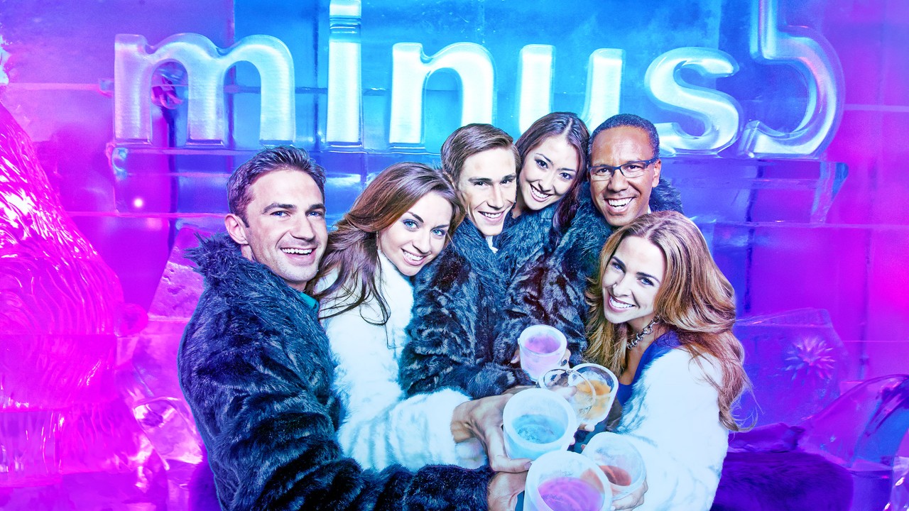 A group of people in warm coats holding drinks with an illuminated "minus5" ice sign in the background.