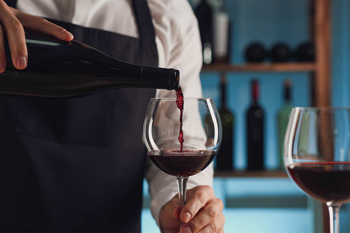 Bartender in a black apron pouring red wine from a bottle into a glass, with another glass of wine nearby.